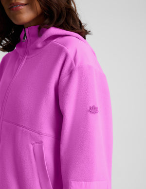 Close-up of a person wearing a bright pink hooded sweatshirt with a partial zipper, pocket, and a small tonal embroidered logo on the upper sleeve, against a light gray studio background.