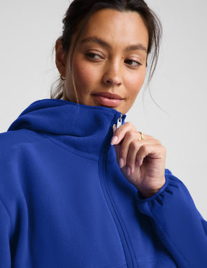 A close-up of a woman wearing a bright royal blue zip-up hoodie, pulling the zipper with her hand against a light gray studio background.