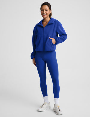 A woman modeling a matching royal blue athletic set consisting of a zip-up hoodie and high-waisted leggings, paired with white socks and white sneakers, against a light gray studio background.