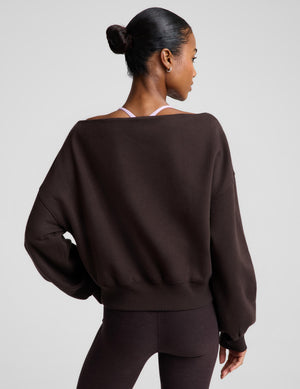 A woman shown from the back wearing a dark brown off-the-shoulder sweatshirt and matching dark brown leggings against a light gray studio background.