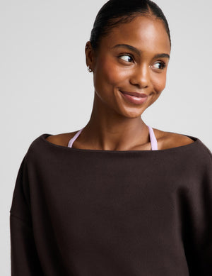 A woman wearing a dark brown off-the-shoulder knit top, photographed against a neutral light gray background.