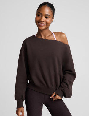 A woman modeling a dark brown off-the-shoulder sweatshirt with balloon sleeves, paired with matching dark leggings, against a solid light gray background.