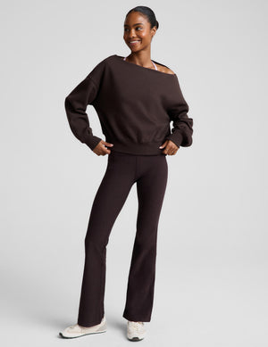 A woman modeling a dark brown off-the-shoulder sweatshirt and matching dark brown flared pants against a light gray studio background.