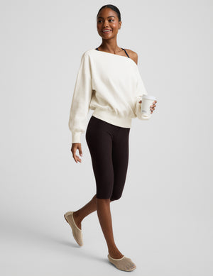 Woman wearing a white off the shoulder fleece sweatshirt and brown leggings against a plain background. 