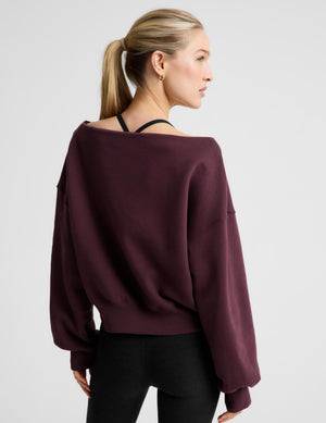 A woman wearing a dark burgundy off-the-shoulder long-sleeve sweatshirt and black pants, photographed against a light gray studio background.