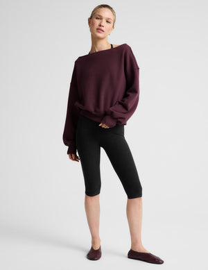 A woman wearing a dark burgundy off-the-shoulder long-sleeve sweatshirt and black pants, photographed against a light gray studio background.