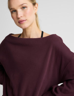A woman wearing a dark burgundy off-the-shoulder long-sleeve sweatshirt and black pants, photographed against a light gray studio background.