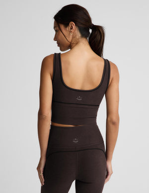 A woman modeling a dark brown activewear set consisting of a scoop-neck crop top and matching high-waisted leggings, photographed against a solid light gray background.