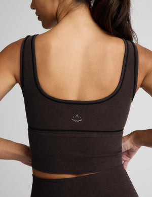 A woman modeling a dark brown activewear set consisting of a scoop-neck crop top and matching high-waisted leggings, photographed against a solid light gray background.