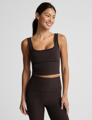 A woman modeling a dark brown activewear set consisting of a scoop-neck crop top and matching high-waisted leggings, photographed against a solid light gray background.