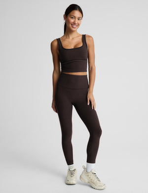 A woman modeling a dark brown activewear set consisting of a scoop-neck crop top and matching high-waisted leggings, photographed against a solid light gray background.