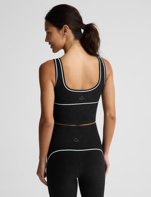 A woman modeling a black athletic crop top with white piping trim and matching black leggings against a light gray studio background.