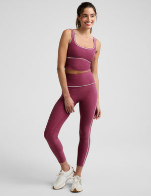 A woman modeling a matching athletic set consisting of a heathered magenta crop top and leggings with thin white contrast piping, photographed against a light gray studio background.