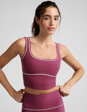 A woman modeling a matching athletic set consisting of a heathered magenta crop top and leggings with thin white contrast piping, photographed against a light gray studio background.