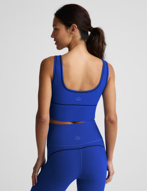 A woman modeling a matching royal blue activewear set consisting of a scoop-neck crop top and high-waisted leggings, photographed against a solid light gray studio background.