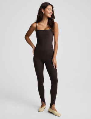 A woman modeling a dark brown, sleeveless, full-length stirrup bodysuit or jumpsuit against a light gray studio background.