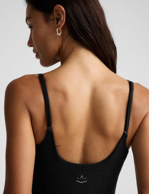 A woman modeling a black sleeveless bodysuit with thin shoulder straps and full-length leggings, paired with black flats, against a light gray studio background.