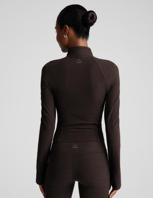 Rear view of a model wearing a dark brown long-sleeve athletic top and matching leggings against a light gray studio background.