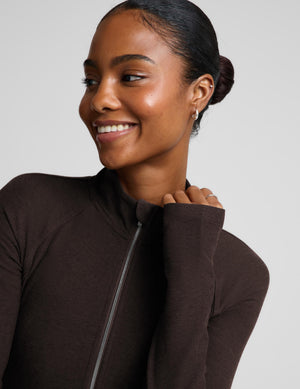A smiling woman with her hair in a bun wearing a dark brown long-sleeve zip-up athletic top, shown in a close-up profile shot against a light gray background.