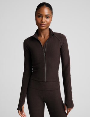 A woman modeling a dark brown long-sleeve athletic zip-up jacket with a stand collar and matching dark brown leggings against a light gray studio background.