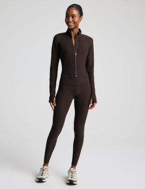 A woman modeling a dark brown jacket and matching high-waisted leggings, paired with white sneakers, against a light gray studio background.