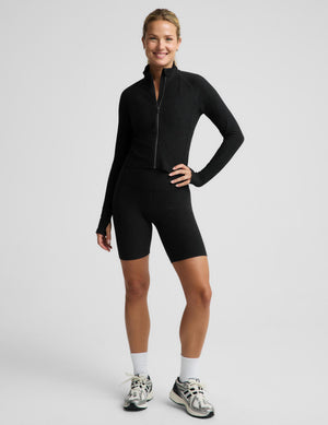 A woman modeling a black long-sleeve athletic zip top and matching black biker shorts against a light gray studio background.