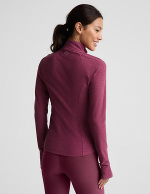 A woman wearing a heathered burgundy long-sleeve athletic quarter-zip fleece and matching leggings, shown from a three-quarters back view against a light gray studio background.