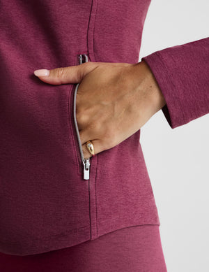 Close-up of a person wearing a heathered burgundy zip-up athletic jacket, showing a hand sliding into a side pocket against a light gray background.