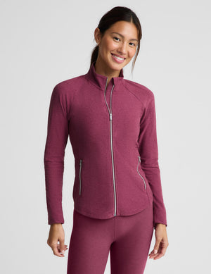 A woman modeling a heathered burgundy athletic zip-up jacket and matching leggings against a light gray studio background.