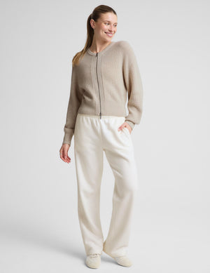 Woman wearing a neutral zip cardigan and white sweatpants against a plain background. 