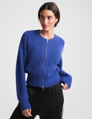 Woman wearing a blue zip cardigan and black track pants against a plain background. 