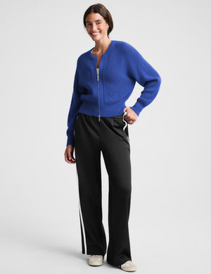 Woman wearing a blue zip cardigan and black track pants against a plain background. 