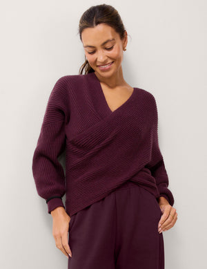 A woman wearing a ribbed knit wrap-style sweater in deep plum purple, paired with matching wide-leg trousers, standing against a light gray studio background.