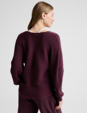 A woman wearing a ribbed knit wrap-style sweater in deep plum purple, paired with matching wide-leg trousers, standing against a light gray studio background.