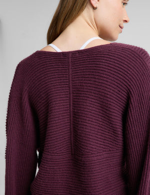 A woman wearing a ribbed knit wrap-style sweater in deep plum purple, paired with matching wide-leg trousers, standing against a light gray studio background.
