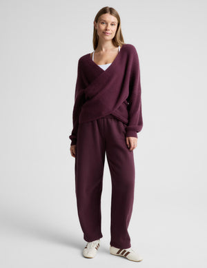 A woman wearing a ribbed knit wrap-style sweater in deep plum purple, paired with matching wide-leg trousers, standing against a light gray studio background.