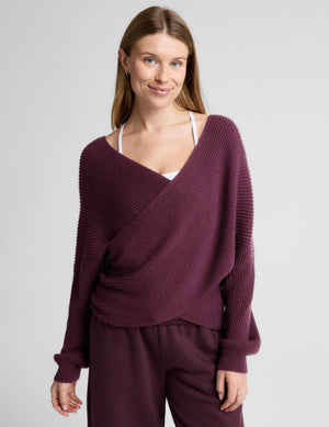 A woman wearing a ribbed knit wrap-style sweater in deep plum purple, paired with matching wide-leg trousers, standing against a light gray studio background.
