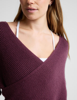 A woman wearing a ribbed knit wrap-style sweater in deep plum purple, paired with matching wide-leg trousers, standing against a light gray studio background.