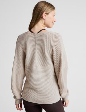 A woman modeling a beige ribbed knit wrap-style sweater with long sleeves, worn over a dark brown camisole and dark leggings, against a light gray studio background.