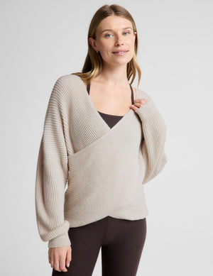 A woman modeling a beige ribbed knit wrap-style sweater with long sleeves, worn over a dark brown camisole and dark leggings, against a light gray studio background.
