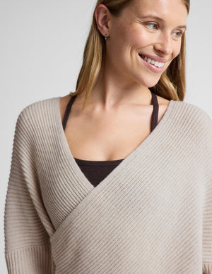 A close-up of a woman wearing a light beige ribbed knit wrap sweater over a dark brown camisole, set against a plain light gray background.