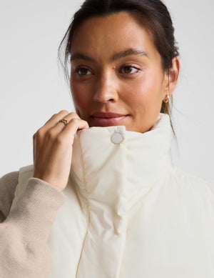 A woman wearing a cream-colored sleeveless puffer vest with beige knit sleeves and dark brown leggings, photographed against a light gray studio background.