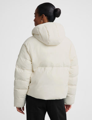 A woman shown from the back wearing a cream-colored puffer jacket with a padded hood and horizontal quilting, paired with black pants, against a plain light gray studio background.