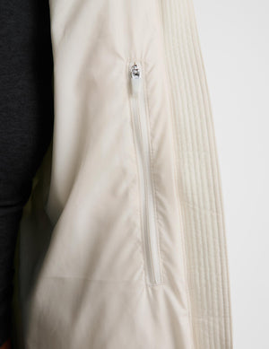 Close-up of a light cream or off-white puffer vest with a vertical zippered pocket, shown on a model wearing dark gray leggings against a plain light background.