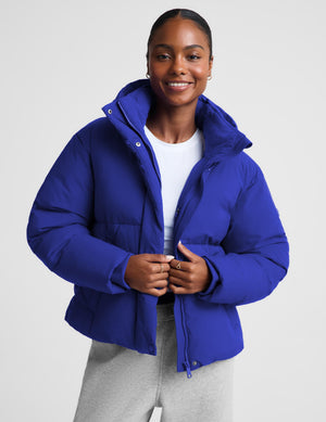 A woman wearing a bright royal blue cropped puffer jacket with a high collar, styled over a white t-shirt and light gray sweatpants, against a light gray studio background.