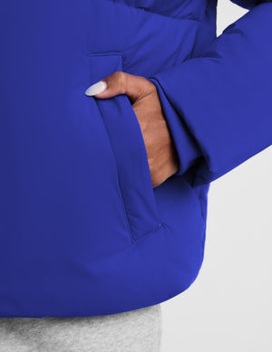 Close-up of a person wearing a bright royal blue puffer jacket, showing a hand with white nail polish inserted into a side pocket, against a light gray background.