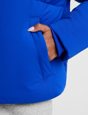 Close-up of a person wearing a bright royal blue puffer jacket, showing a hand with white nail polish sliding into a side pocket.