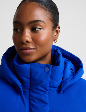 Close-up of a model wearing a bright royal blue puffer jacket with a high padded collar and snap-button placket, against a light gray studio background.