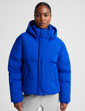 A woman wearing a bright royal blue hooded puffer jacket with horizontal quilting, paired with light gray sweatpants, photographed against a plain light gray studio background.