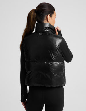 A woman modeling a black puffer vest with a shiny, quilted texture over a black long-sleeve top and black leggings, set against a solid light gray background.